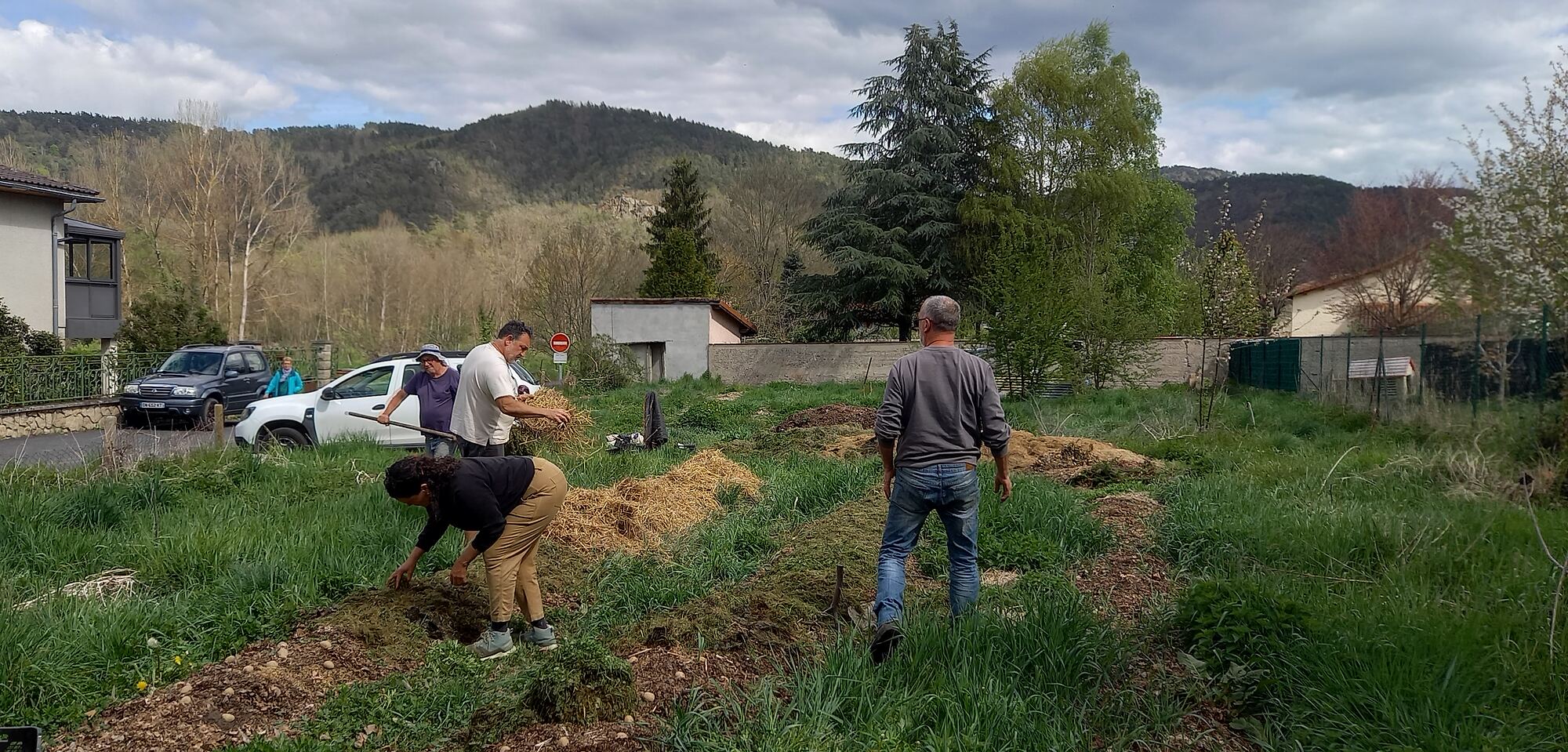 Quatre personnes étalent de la paille et du compost sur des buttes de culture, devant des montagnes boisées.