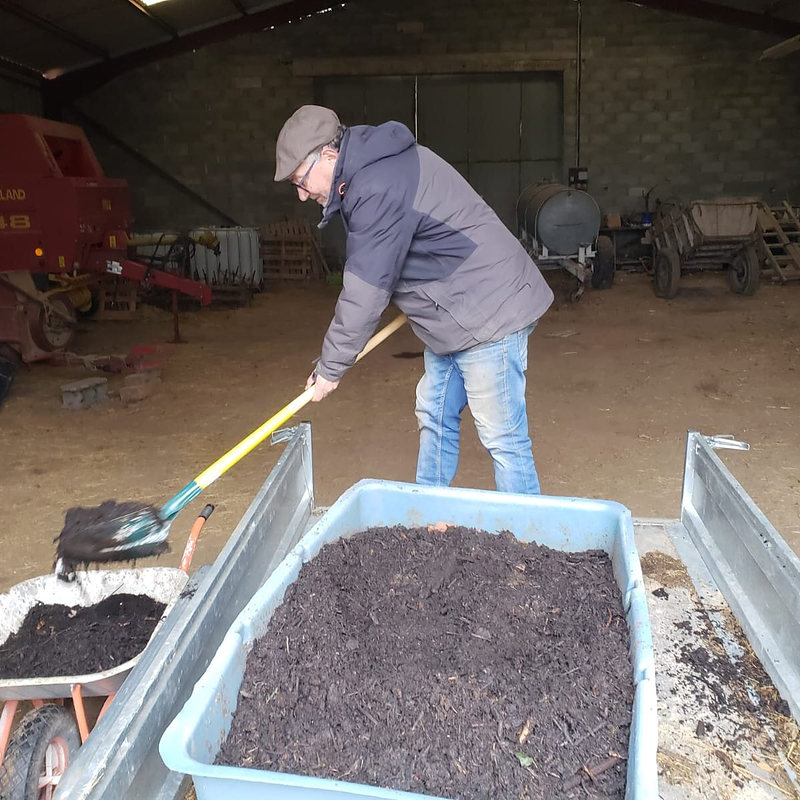 Une personne charge du compost à la pelle dans une remorque, sous un hangar.