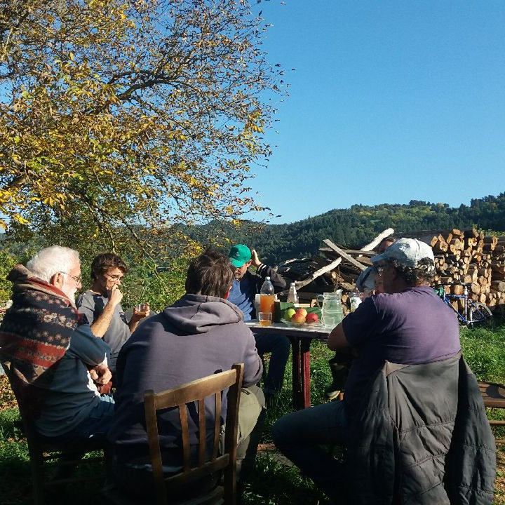 Six personnes attablées en plein air sous un arbre, devant des collines boisées.